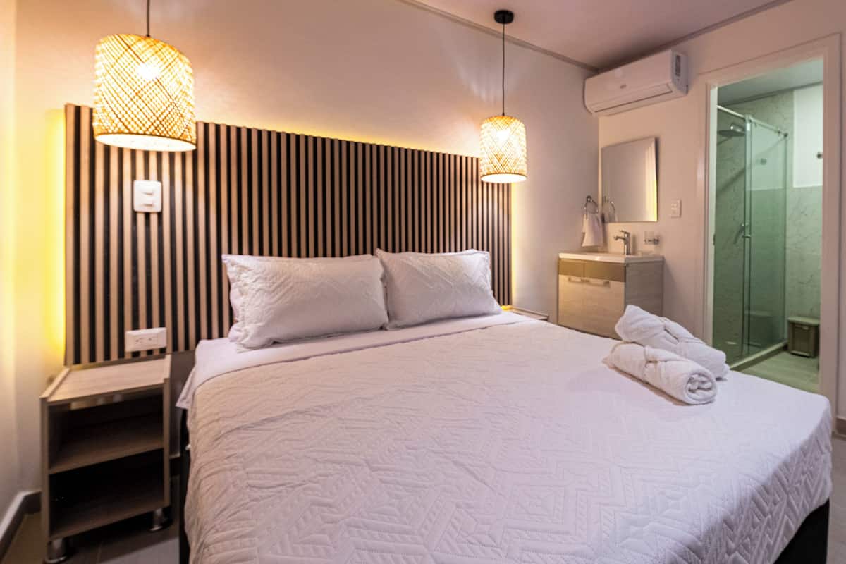 A hotel room with a bed, bedside lamps, a vanity with a mirror, and a shower area.