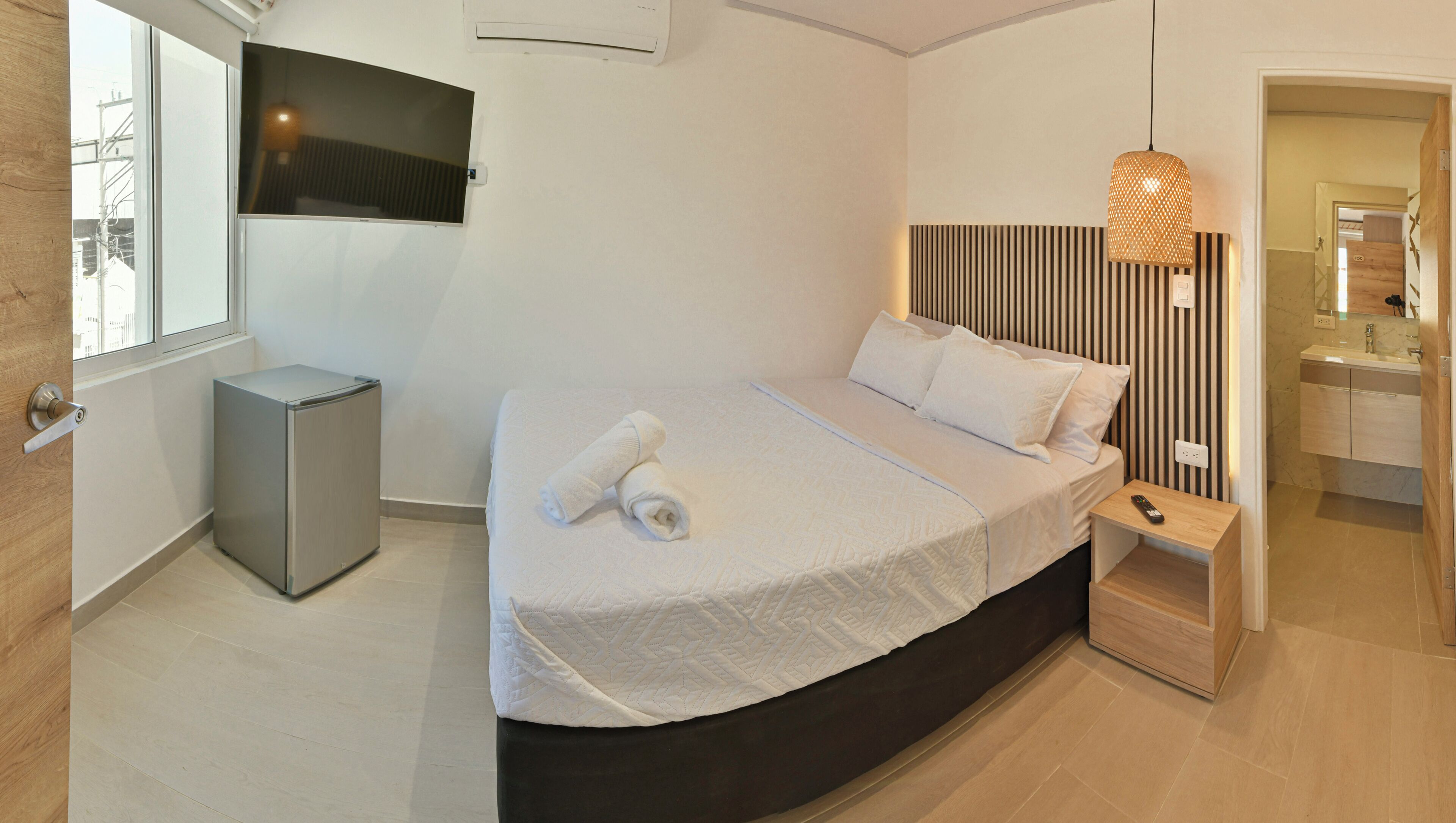 A modern hotel room with a large bed, a bedside table, a TV mounted on the wall, and a bathroom visible through an open door.