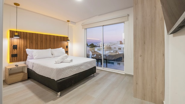 A modern bedroom with a large bed, wooden headboard, bedside tables, and a view of buildings through a sliding glass door. - Hotel Blu Sai (San Andres)
