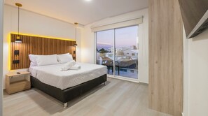 A modern bedroom with a large bed, wooden headboard, bedside tables, and a view of buildings through a sliding glass door. - Hotel Blu Sai (San Andres)