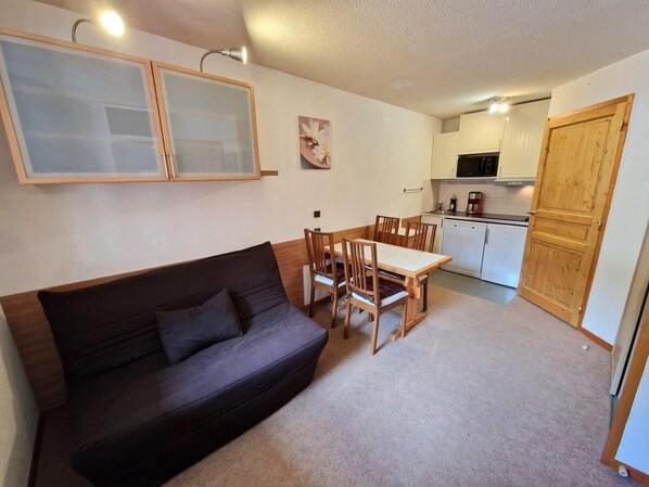 A compact living space with a dark-colored sofa, a dining table with chairs, a kitchenette with cabinets, and a wooden door.