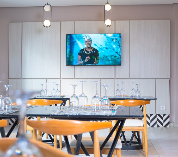 A modern dining area with wooden tables and chairs, a flat-screen TV mounted on the wall, and a decorative wall panel. - Manou Hotel (Douala)