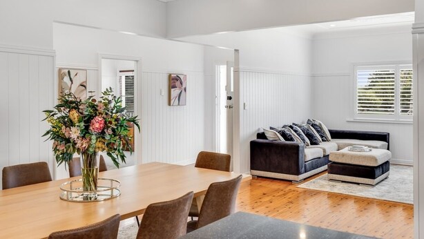 A spacious living area with a wooden dining table, a dark sofa set, and a large window with blinds. - Botanic Breeze on Lindsay! (East Toowoomba)