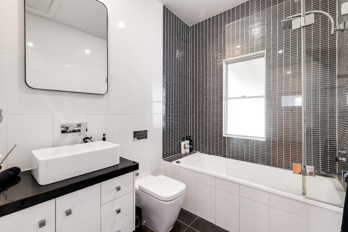 Et moderne badeværelse med en hvid vask, bordplade i sort, et hvidt toilet og et badekar med brusekabine i glas.