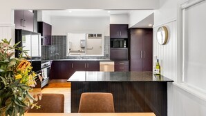 A modern kitchen with dark cabinets, a central island, and a dining area with a table and chairs. - Botanic Breeze on Lindsay! (East Toowoomba)