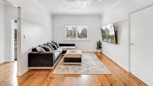 A spacious living room with a dark-colored sofa, a matching ottoman, and a large flat-screen TV mounted on the wall. - Botanic Breeze on Lindsay! (East Toowoomba)
