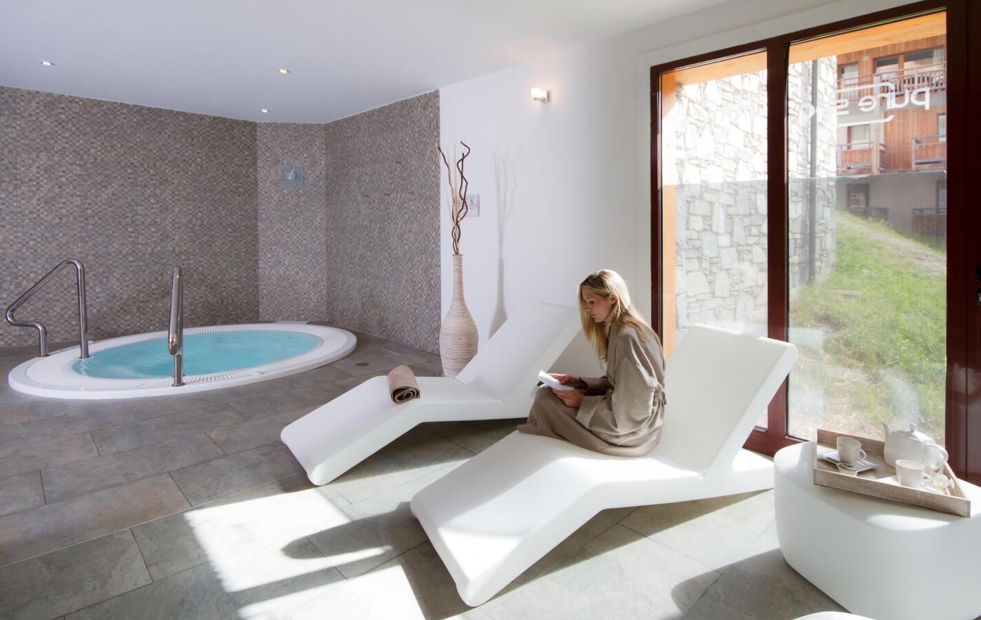 Une salle de spa moderne équipée d’un jacuzzi, d’un fauteuil de relaxation et d’une grande fenêtre donnant sur un bâtiment en pierre.