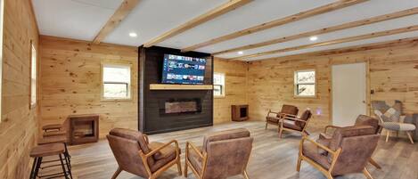 Een ruime kamer met houten wanden en een grote flatscreen televisie bevestigd aan een donkere muur. Er staan verschillende leren fauteuils rond een centraal gebied, en een klein houten tafeltje met krukjes.