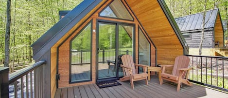 A cabin with a sloped roof, a deck with wooden chairs, and a glass door leading to an outdoor area.