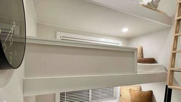 A compact room with a bunk bed, a wall clock, an air conditioning unit, and a window with blinds.