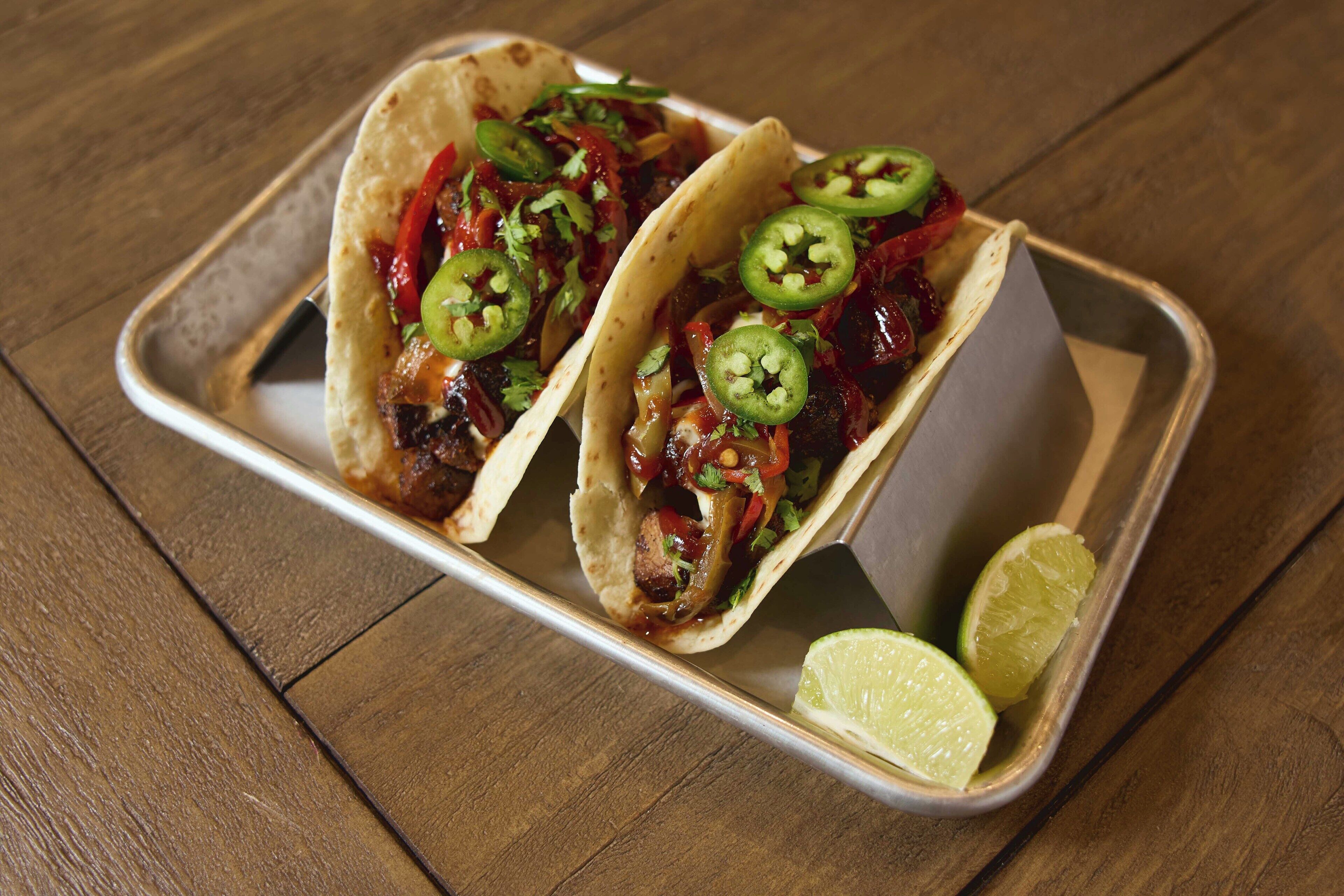 Deux tacos garnis de jalapeños et accompagnés de quartiers de citron vert, servis sur un plateau en métal.