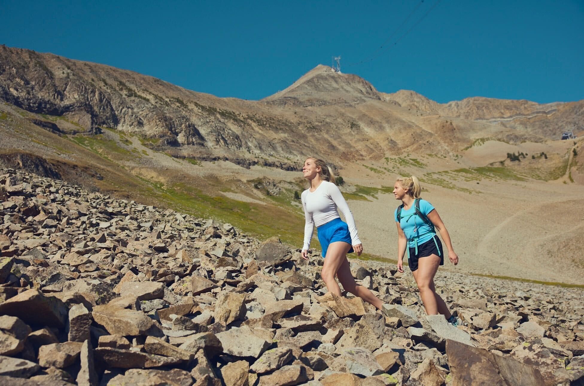 Twee mensen wandelen over rotsachtig terrein, met een berg op de achtergrond.