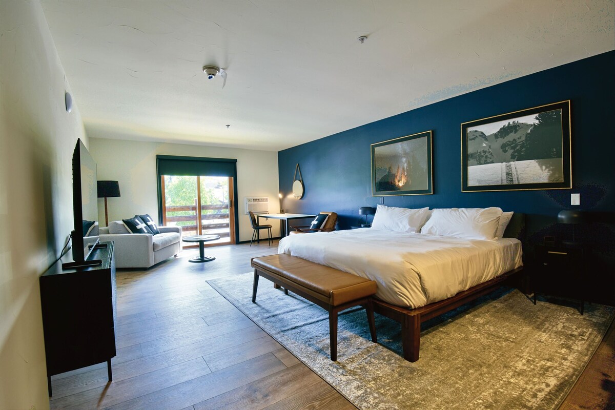 A modern bedroom with a large bed, a desk, a sofa, and a view of the outdoors.