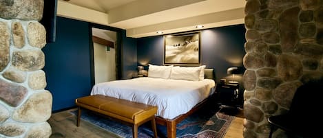 A bedroom with a stone wall, a bed with white linens, a wooden bench, and a rug.