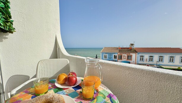 A balcony with a table set for breakfast, including a donut, fruit, and juice, overlooking a coastal town and the sea.