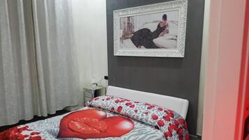 A bedroom with a bed, a nightstand, a framed picture on the wall, and a window with curtains.
