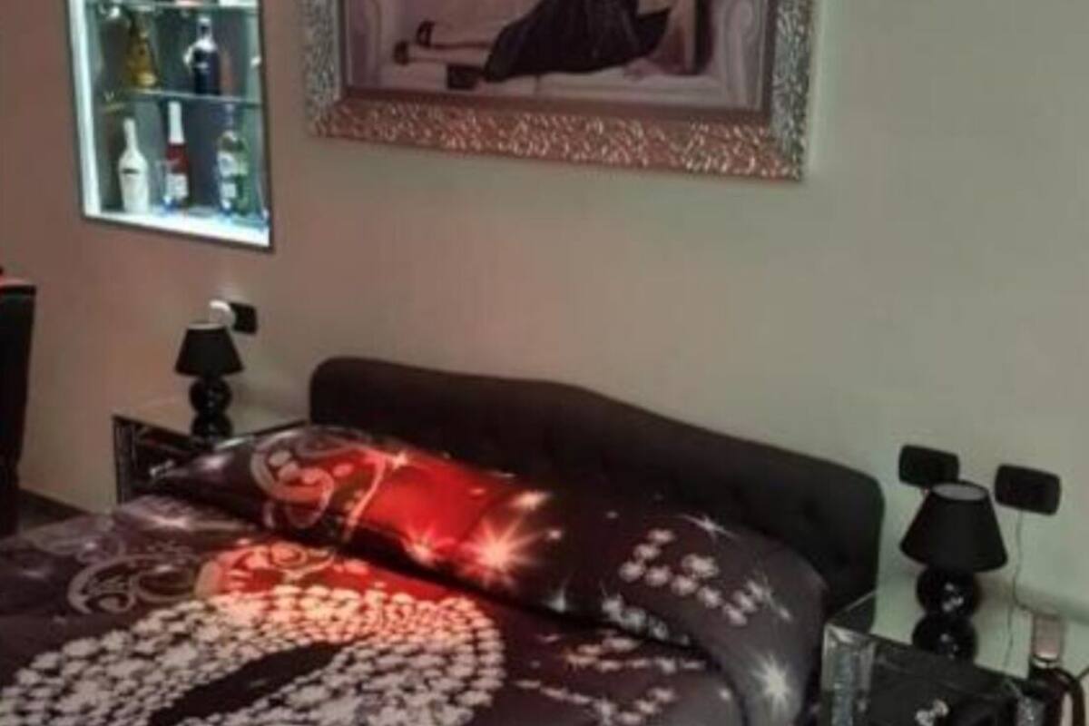 Ein Schlafzimmer mit einem Bett, einem Nachttisch, einer Lampe, einem Regal mit Flaschen und einem gerahmten Bild an der Wand.