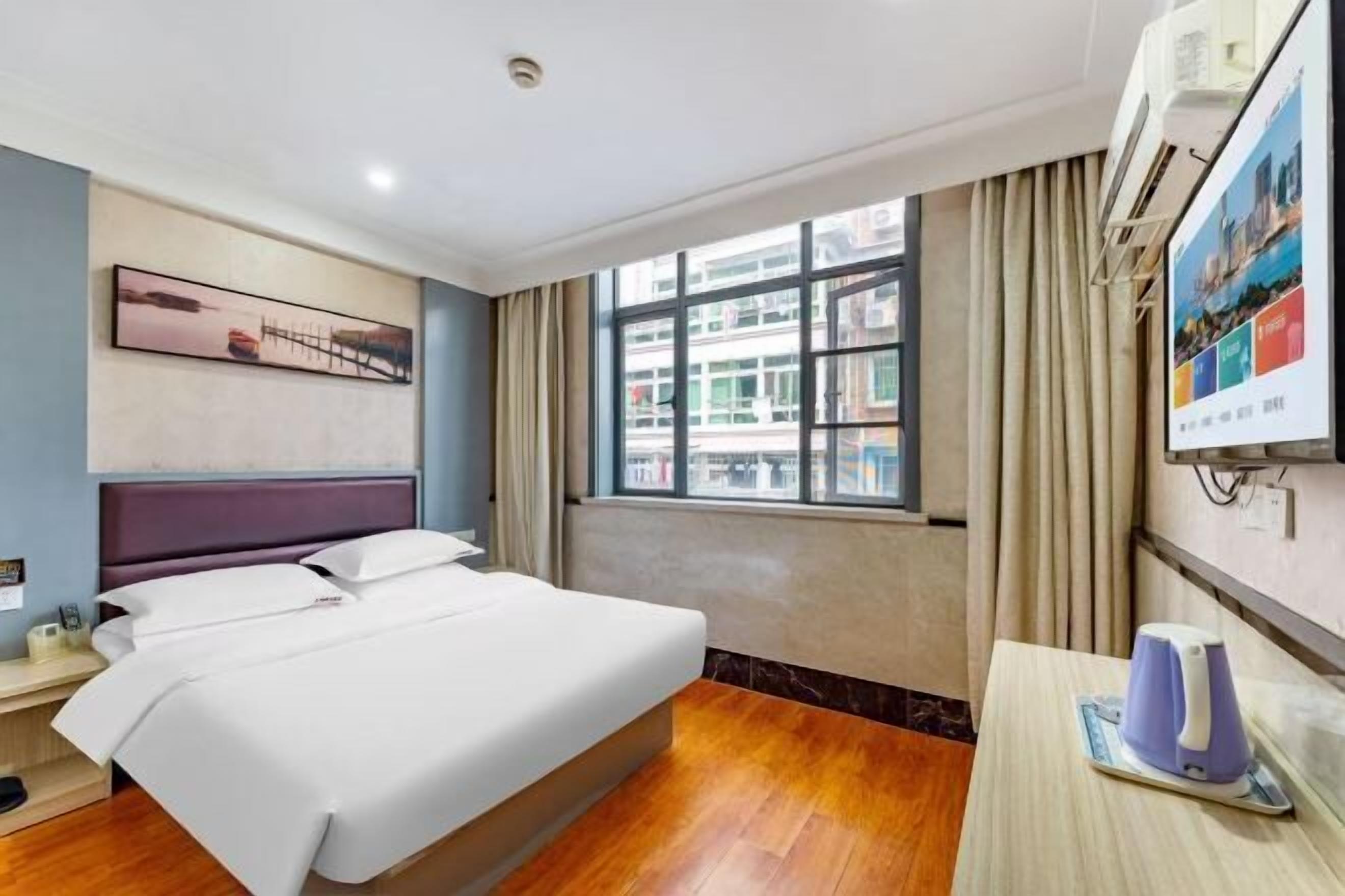 A hotel room with a bed, a desk, a kettle, and a window with curtains.