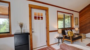 A room with a wooden-framed door, a black cabinet, a sofa with a cushion, a lamp, and a window with a view of greenery.