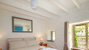 A bedroom with a bed, a bedside table, a lamp, a mirror, and a window with curtains. - Hollies Cottage (Truro)