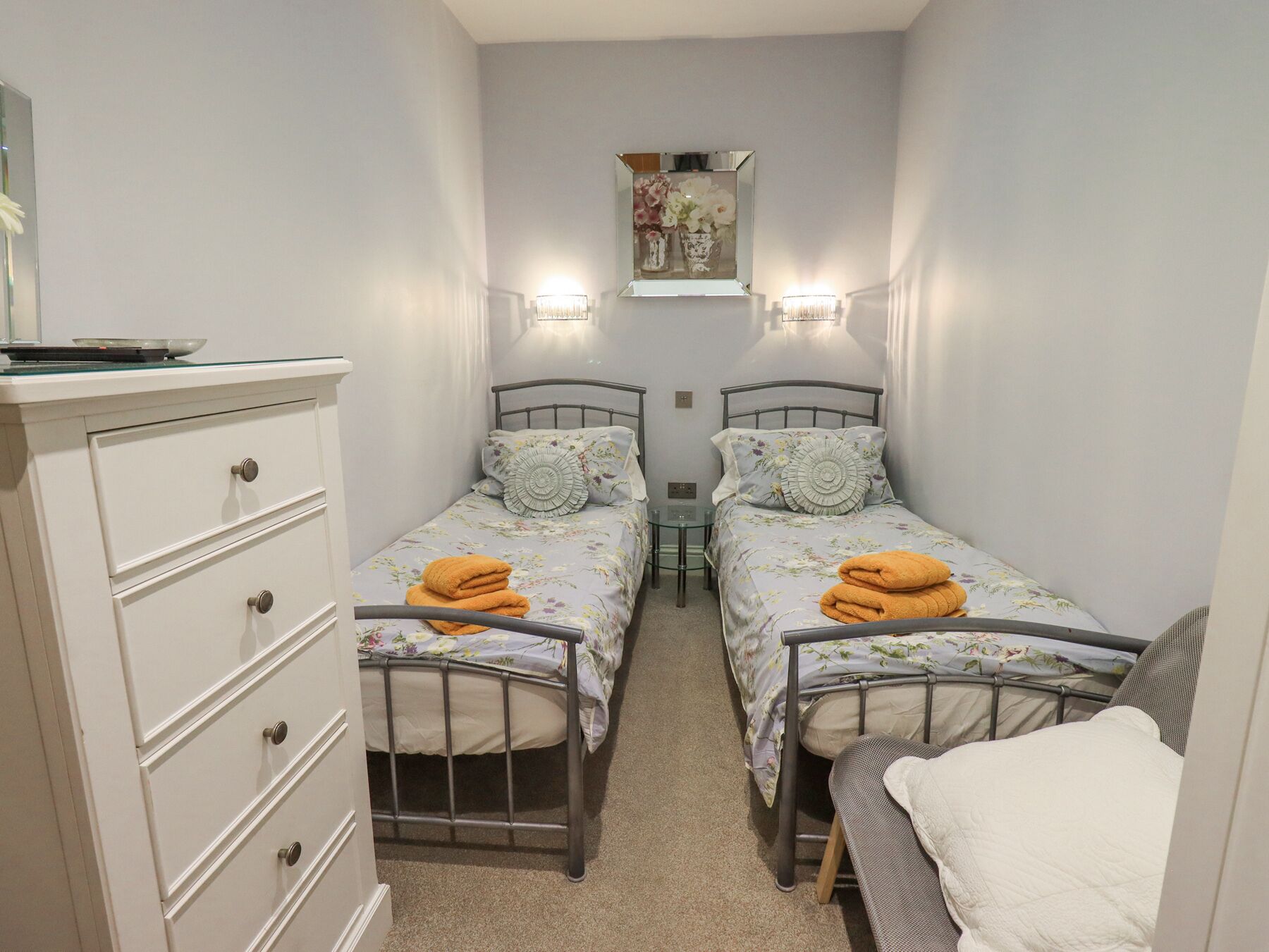 A narrow room with two single beds, each with a floral bedspread and a stack of yellow towels. There is a white dresser with drawers and a mirror above it.
