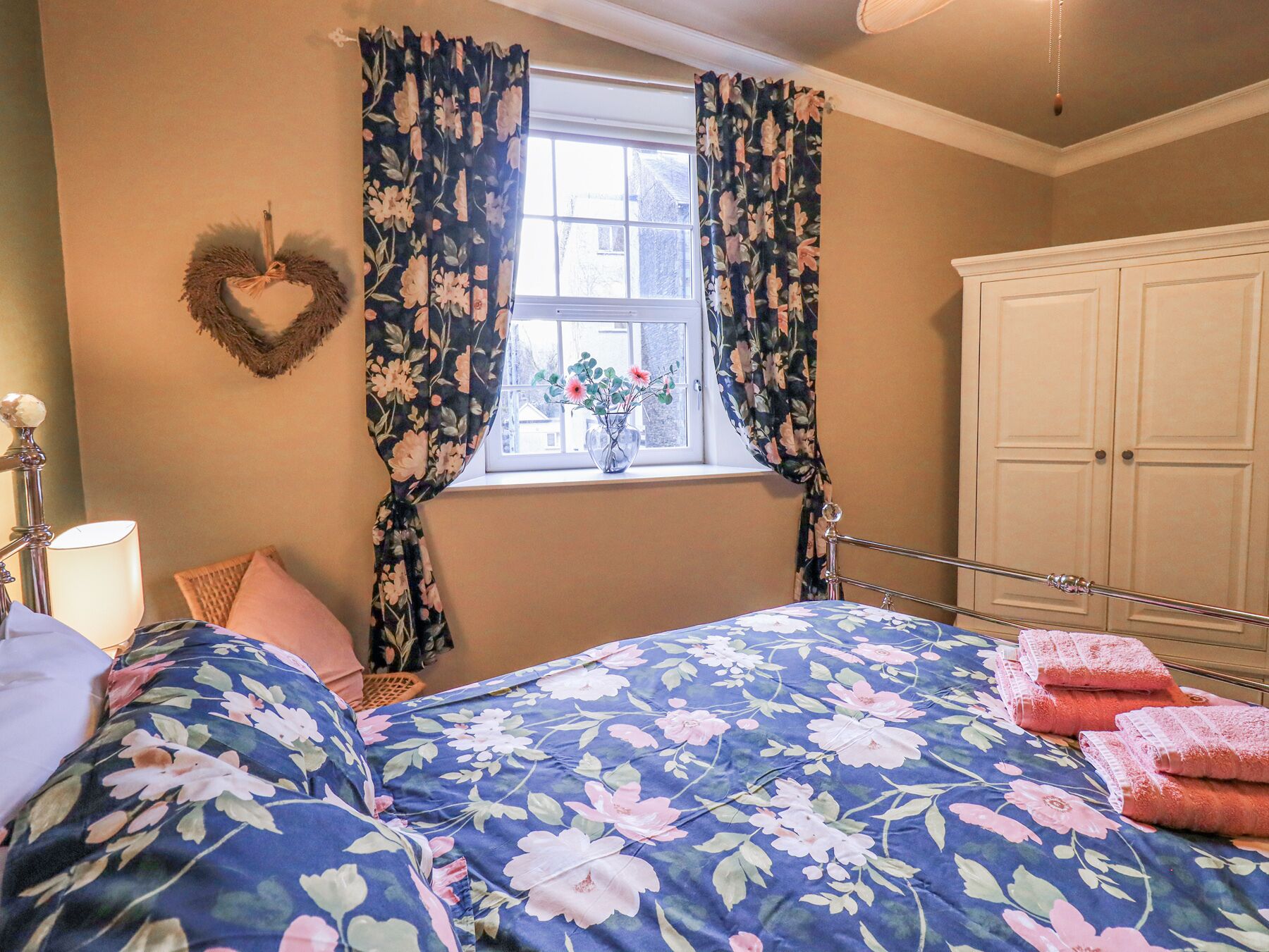 Une chambre à coucher avec une couvre-lit à motifs floraux, une décoration en forme de cœur accrochée au mur et une fenêtre avec des rideaux.