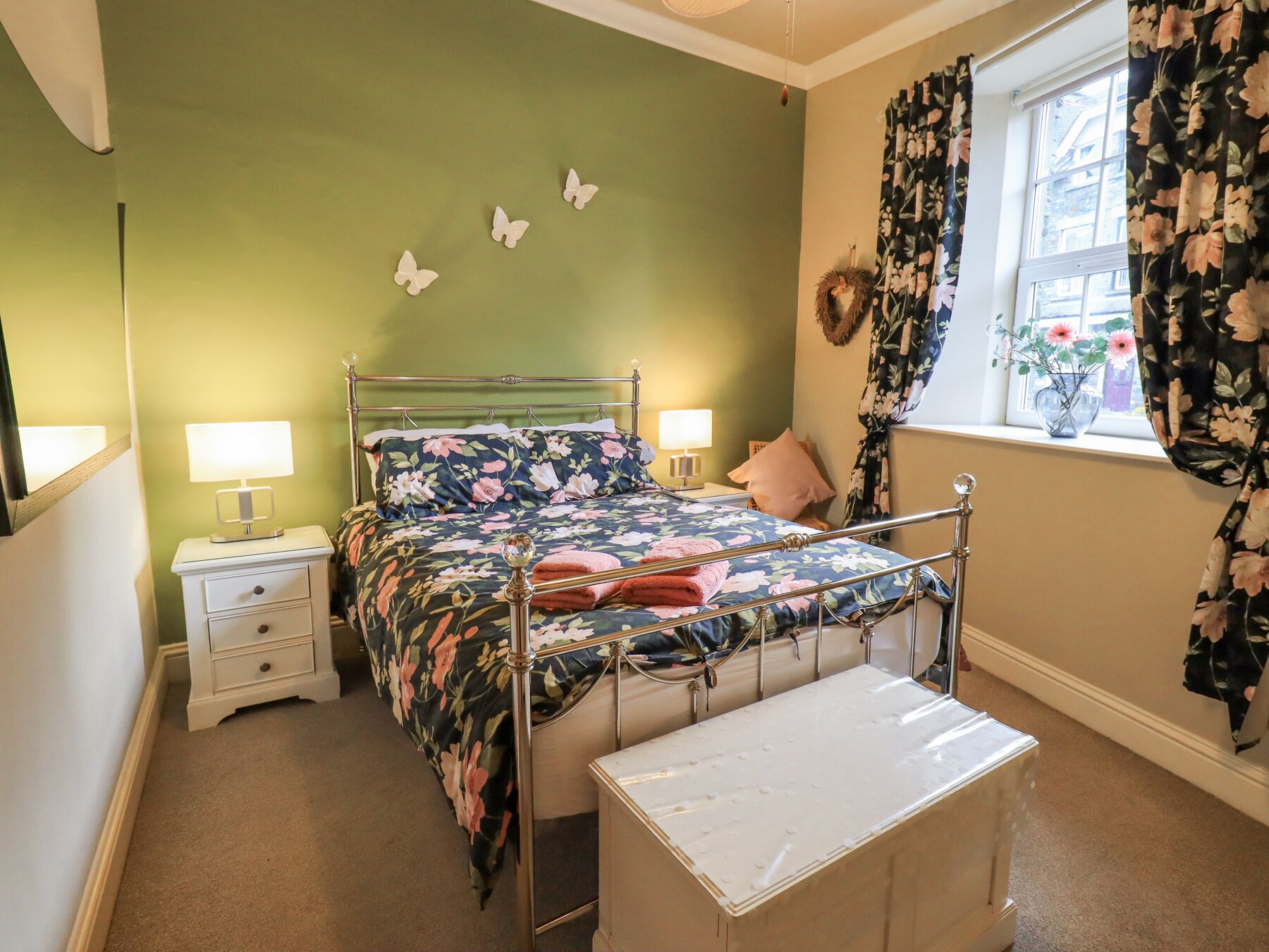 A bedroom with a metal bed frame, floral bedding, a bedside table with a lamp, a window with floral curtains, and a small wall decoration.