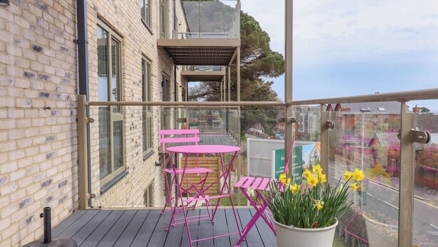 A balcony with a wooden floor, a pink metal table and chairs, and a potted plant with yellow flowers. - Rolles Rainbow (Exmouth)