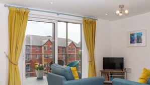 A modern living room with a large sliding glass door, two blue sofas with yellow cushions, a flat-screen TV, and a small side table. - Rolles Rainbow (Exmouth)