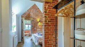 A narrow hallway with a brick wall, a wooden floor, a bed with pillows, a radiator, and a wooden wardrobe with hanging clothes. - Beautiful romantic converted barn located in a quiet setting. (Wingfield, near Diss)