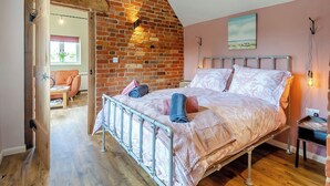 A bedroom with a brick wall, a bed with a metal frame, a nightstand, and a painting on the wall. - Beautiful romantic converted barn located in a quiet setting. (Wingfield, near Diss)