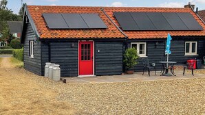 A wooden house with a red door, solar panels on the roof, and a patio with outdoor furniture. - Beautiful romantic converted barn located in a quiet setting. (Wingfield, near Diss)