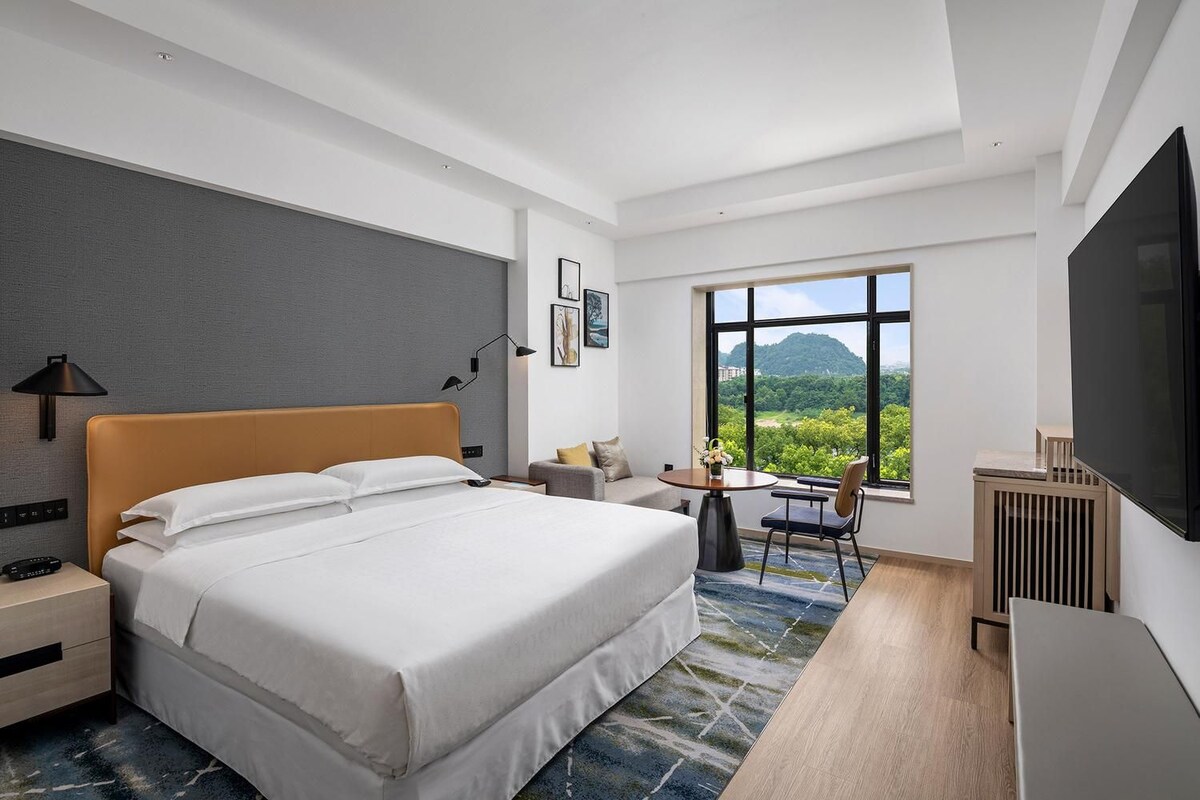 A modern hotel room with a large bed, a desk, a chair, a TV, and a window with a view of greenery.