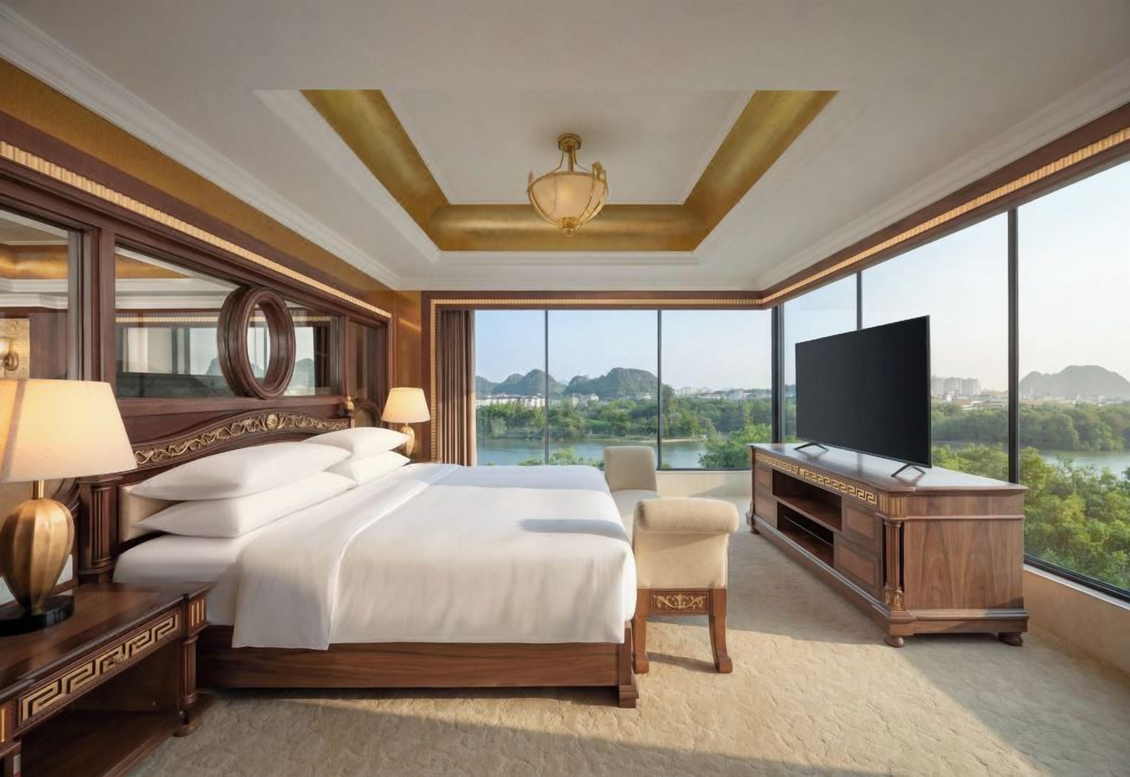 A spacious hotel room with a large bed, a television, a seating area, and a view of a cityscape through large windows.