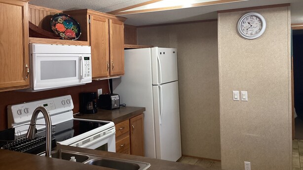Une cuisine de taille réduite avec des meubles en bois, un micro-ondes blanc, un réfrigérateur blanc, une cuisinière et une horloge au mur.