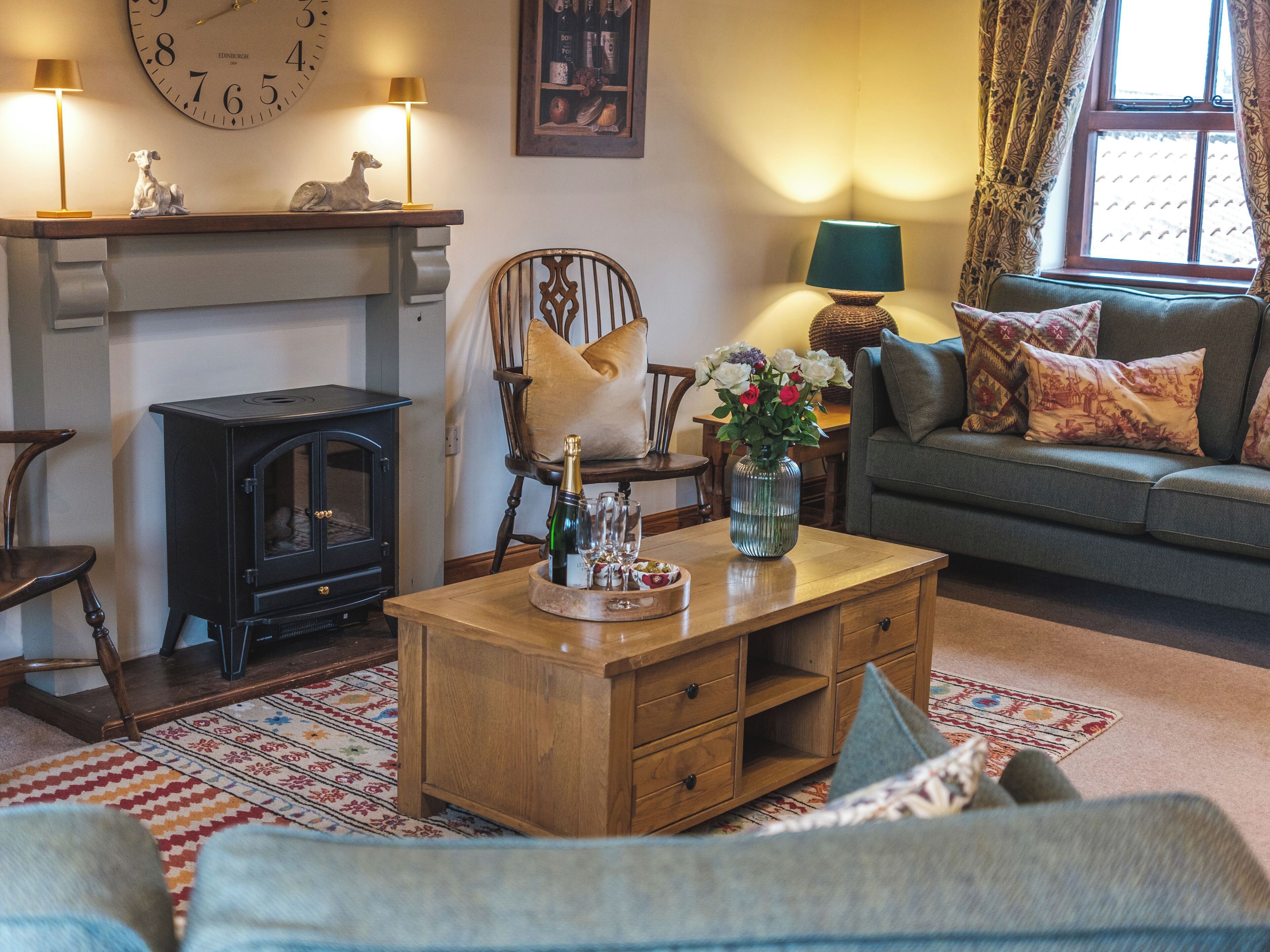 A cozy living room with a fireplace, a sofa, a wooden coffee table with a vase of flowers, and a patterned rug.