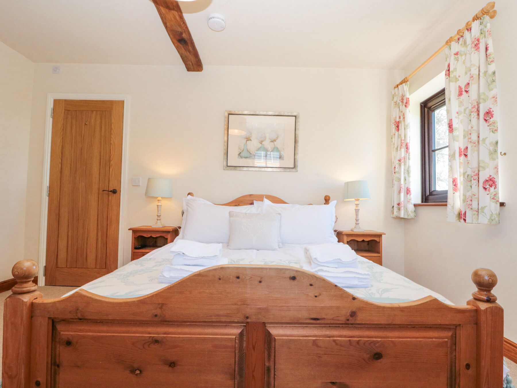A neatly made bed with a wooden headboard, two bedside tables with lamps, a framed picture above the bed, and a window with floral curtains.