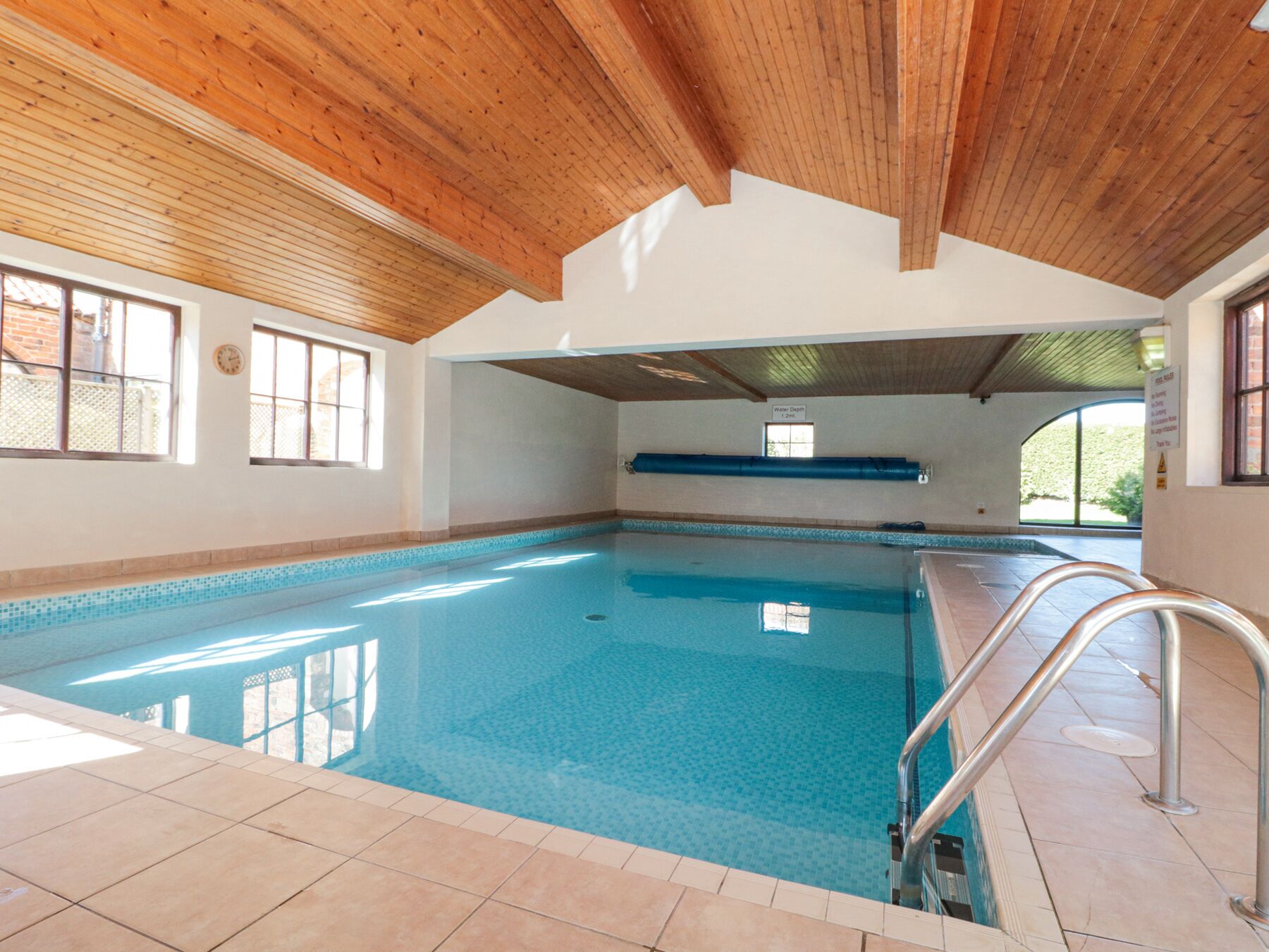 Une piscine intérieure avec un plafond en bois, un sol carrelé et une eau d’un bleu limpide.