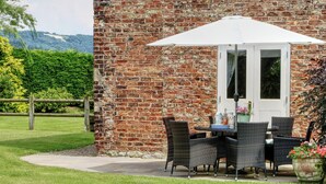 A brick building with a patio area featuring wicker furniture and a white umbrella. - Beaver Cottage (Thirsk)