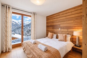 Un dormitorio con cabecera de madera, cama con ropa de cama blanca y vistas a montañas nevadas, además de una ventana con cortinas.
