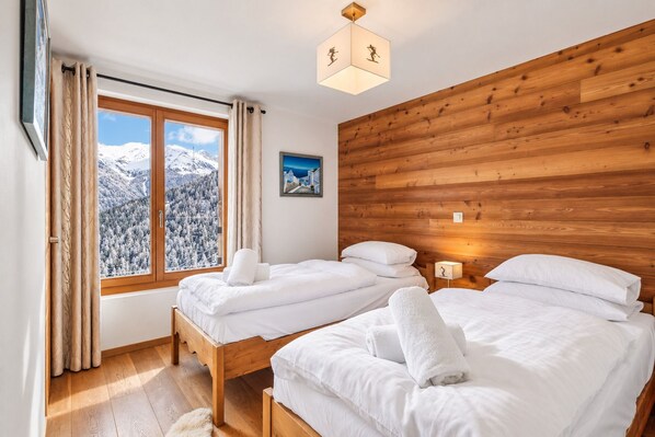Un dormitorio con dos camas, paneles de madera en la pared, un ventanal con vistas a una montaña nevada y un cuadro.