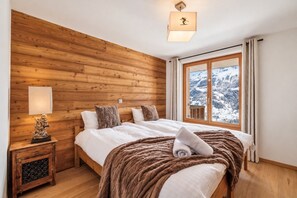 Un dormitorio con pared de madera, una cama con ropa blanca, una manta marrón, una mesita de noche con una lámpara y una ventana con vistas nevadas.