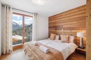 Una camera da letto con una testiera in legno, una grande finestra con vista sulle montagne e un letto con due cuscini e un asciugamano arrotolato.