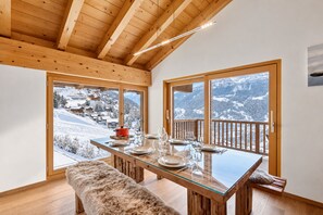 Comedor con mesa de vidrio, banco de madera y vista a un paisaje nevado de montaña.