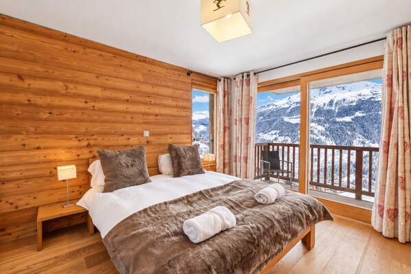 Un dormitorio con pared de madera, una cama grande con un cubrecolcha de piel, una mesita de noche y vistas a montañas nevadas desde un balcón.