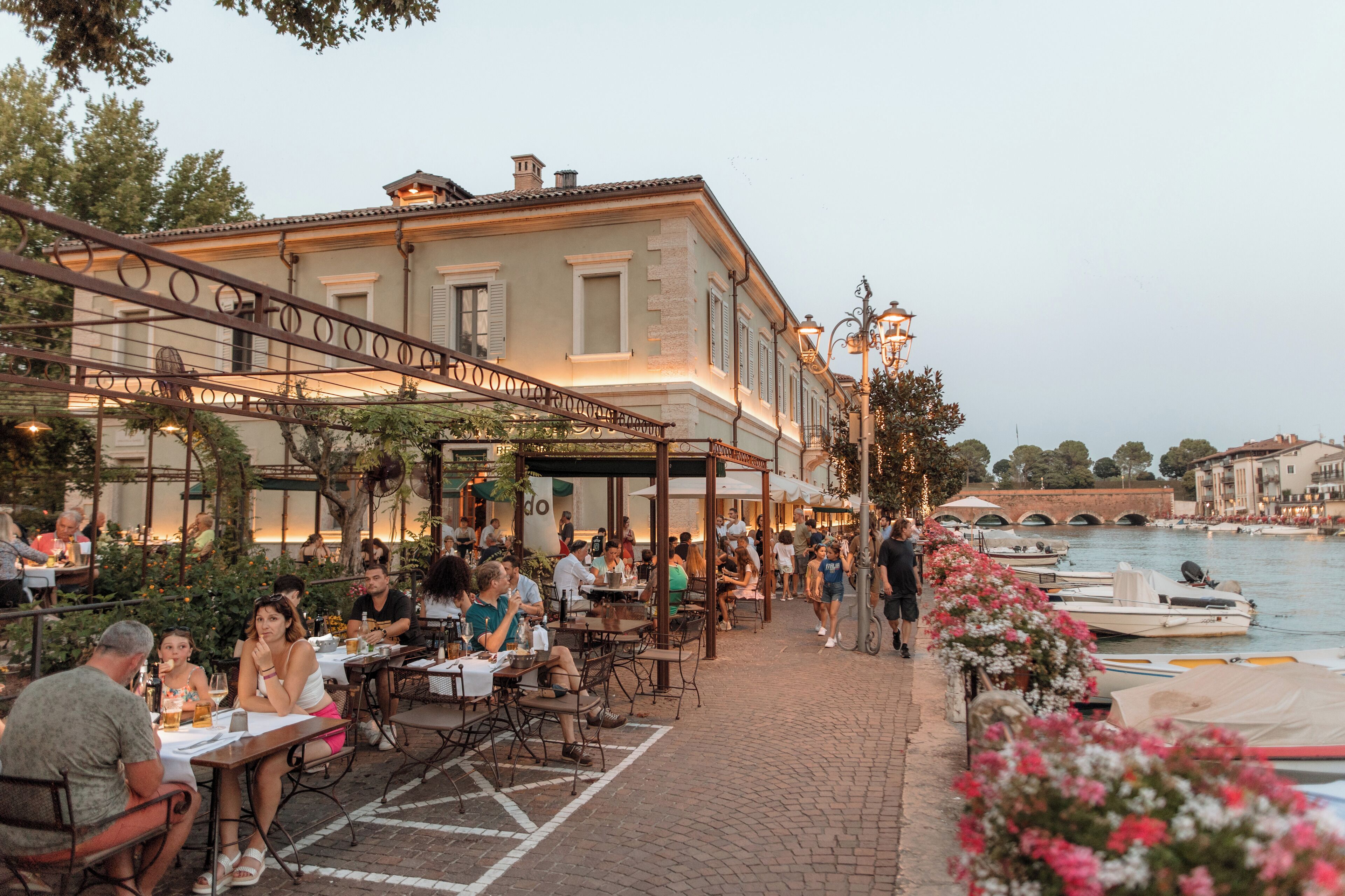 Un área de comedor al aire libre con mesas y sillas, un edificio con balcón y un río con barcos al fondo.