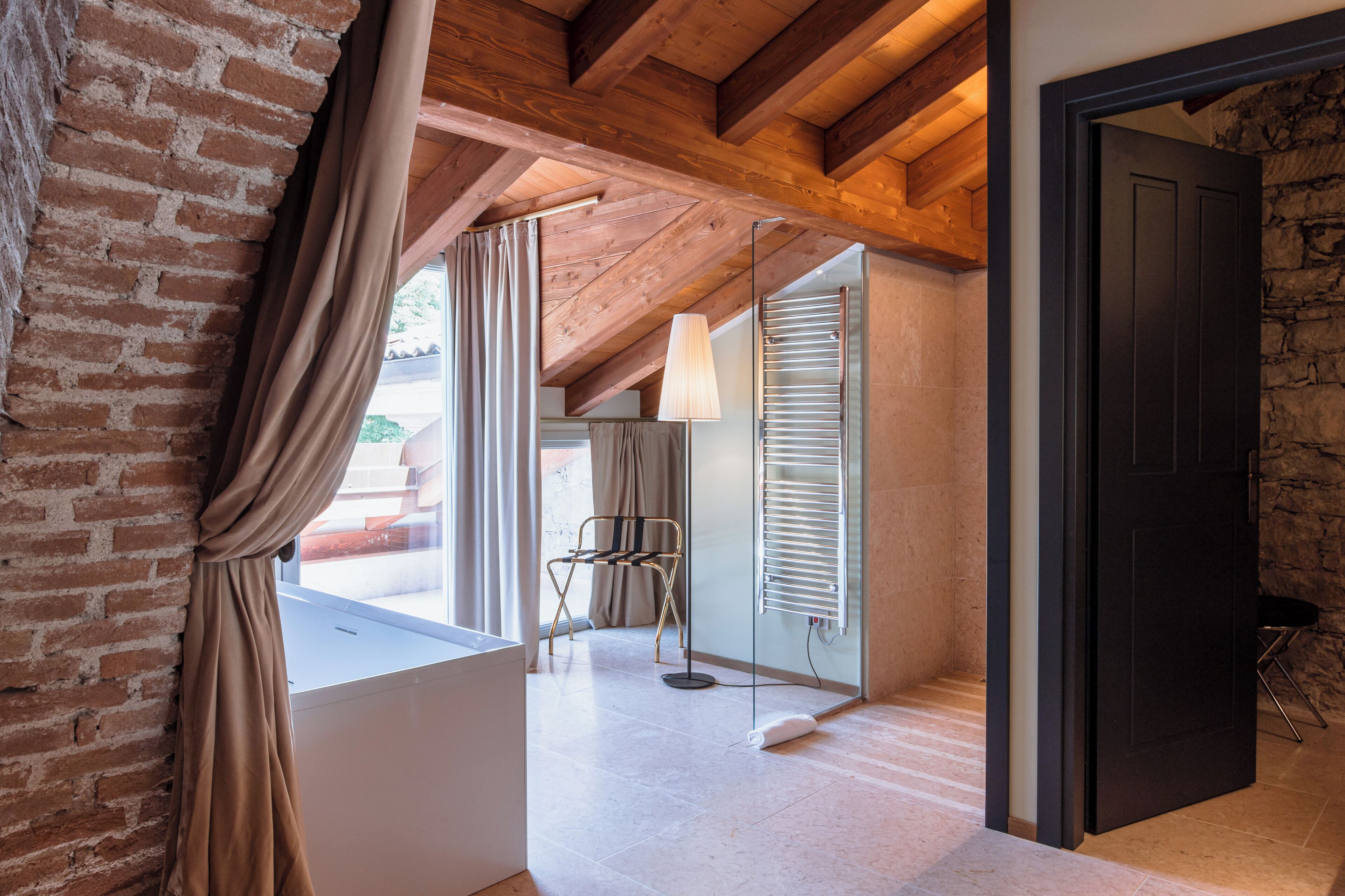 Una habitación con vigas de madera a la vista, pared de ladrillo, ventana con cortinas, una silla y una puerta.