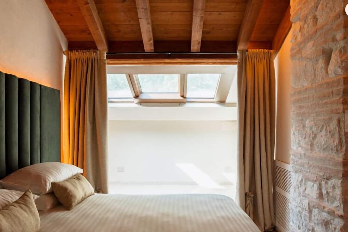 Quarto com cama, cortinas, teto de madeira e vista pela janela.