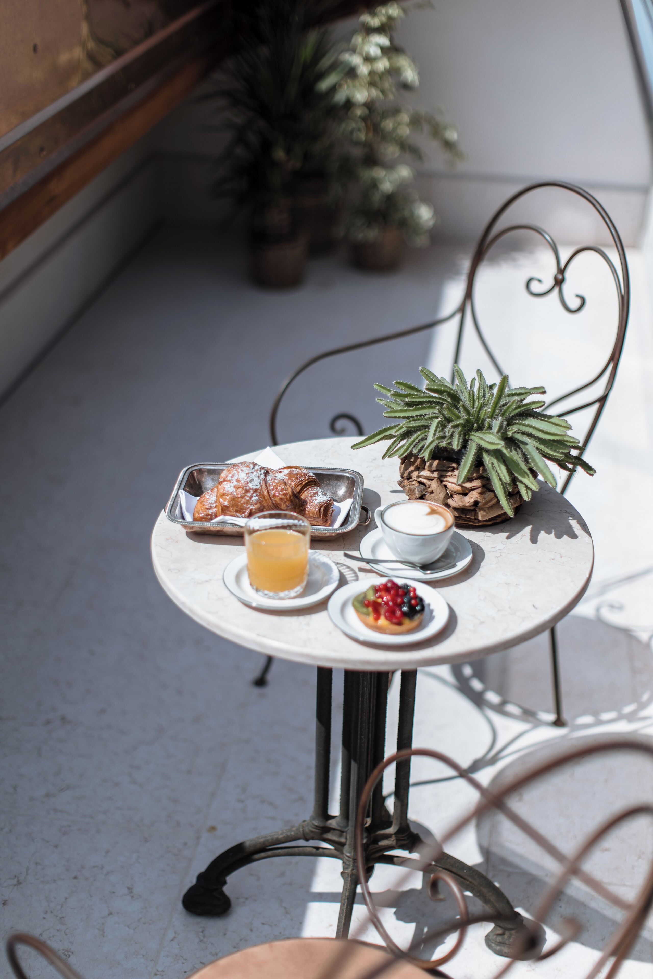 Une petite table ronde avec une chaise en métal, un pot de fleurs, une assiette de pâtisseries, une tasse de café, un verre de jus d’orange et une petite assiette de fruits rouges.