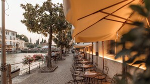 A riverside café with outdoor seating, a cobblestone path, and a view of buildings and a bridge in the background. - Pavillon Suite (Peschiera del Garda)